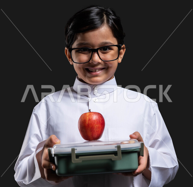 أكل الفواكه الطازجة للشعور بالحيوية والنشاط , إيماءات النظر إلى الكاميرا, تناول وجبة فطور صحية في وقت الفسحة , بورتريه مقرب لولد عربي خليجي سعودي مبتسم  يرتدي الثوب التقليدي والنظارة الطبية يحمل بيده حافظة الطعام المدرسية , العودة إلى المدارس , خلفية سوداء .