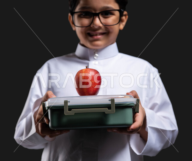 العودة إلى المدارس ,إيماءات النظر إلى الكاميرا, تناول وجبة فطور صحية في وقت الفسحة , بورتريه مقرب لولد عربي خليجي سعودي مبتسم  يرتدي الثوب التقليدي والنظارة الطبية يحمل بيده حافظة الطعام المدرسية , أكل الفواكه الطازجة للشعور بالحيوية والنشاط,  خلفية سوداء .