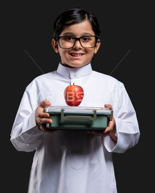 تناول وجبة فطور صحية في وقت الفسحة, إيماءات النظر إلى الكاميرا,  بورتريه مقرب لولد عربي خليجي سعودي مبتسم  يرتدي الثوب التقليدي والنظارة الطبية يحمل بيده حافظة الطعام المدرسية ,أكل الفواكه الطازجة للشعور بالحيوية والنشاط, العودة إلى المدارس , خلفية سوداء .