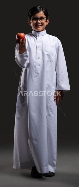 وجبة فطور  صحية طازجة , تناول الطعام الصحي في وقت الفسحة , بورتريه لولد عربي خليجي سعودي مبتسم  يرتدي الثوب التقليدي والنظارة الطبية يحمل بيده تفاحة حمراء وينظر إلى الكاميرا , مفهوم العناية والاهتمام  بالصحة, صورة الجسم بالطول الكامل , خلفية سوداء .