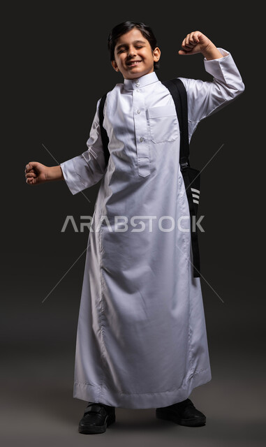 العودة إلى المدارس, الشعور بالكسل والخمول للذهاب باكرا إلى المدرسة, بورتريه لفتى عربي سعودي خليجي يرتدي الثوب المدرسي ويحمل الحقيبة يتثاءب, إيماءات النعاس و الإحساس بالتعب, الحاجة إلى النوم والراحة, صورة الجسم بالطول الكامل, خلفية رمادية .