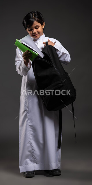 تجهيز المستلزمات المدرسية, الاستعداد للذهاب إلى المدرسة, بورتريه مقرب لولد عربي سعودي خليجي مبتسم يرتدي الثوب المدرسي يقوم بوضع الكتب الدراسية في حقيبته, الشعور بالفرح والحماس في بداية العام الدراسي الجديد, صورة الجسم بالطول الكامل, خلفية رمادية .