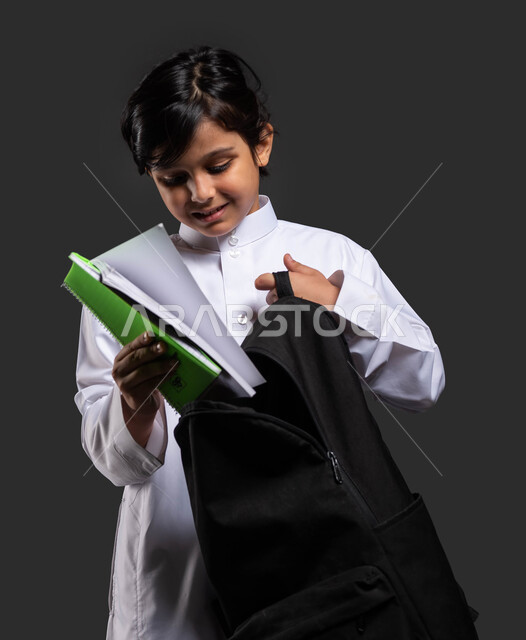 تحضير المستلزمات المدرسية, الاستعداد للذهاب إلى المدرسة, بورتريه مقرب لولد عربي سعودي خليجي مبتسم يرتدي الثوب المدرسي يقوم بوضع الكتب الدراسية في حقيبته, الشعور بالفرح والحماس في بداية العام الدراسي الجديد, التعليم في المملكة العربية السعودية, خلفية سوداء.