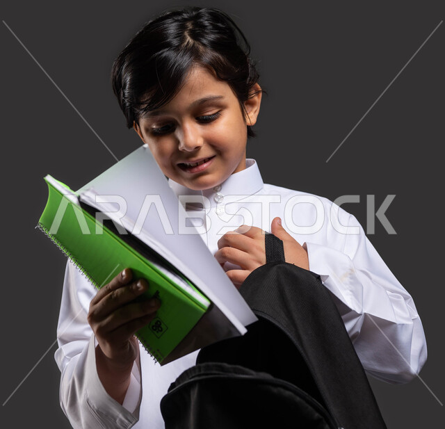 الاستعداد للذهاب إلى المدرسة, بورتريه مقرب لفتى عربي سعودي خليجي مبتسم يرتدي الثوب المدرسي يقوم بوضع الكتب الدراسية في حقيبته, الشعور بالفرح والحماس في بداية العام الدراسي الجديد, التعليم في المملكة العربية السعودية, خلفية سوداء .