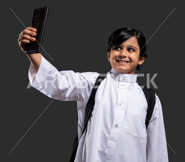 توثيق اللحظات الأولى من العام الدراسي الجديد, استخدام الهاتف المحمول والتقنيات الحديثة, بورتريه مقرب لفتى عربي سعودي خليجي مبتسم يرتدي الثوب التقليدي ويحمل الحقيبة المدرسية يلتقط السيلفي التذكاري, الدراسة والتعليم عن بعد, إيماءات السعادة والفرح, خلفية سوداء .