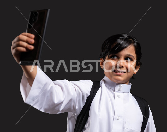 استخدام الهاتف المحمول والتقنيات الحديثة, بورتريه مقرب لفتى عربي سعودي خليجي مبتسم يرتدي الثوب التقليدي ويحمل الحقيبة المدرسية يلتقط السيلفي التذكاري, توثيق اللحظات الأولى من العام الدراسي الجديد , إيماءات السعادة والفرح بالعودة إلى المدرسة, خلفية سوداء.