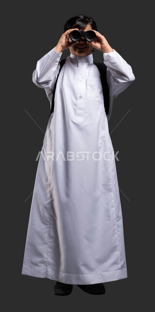 Educational activities to develop the mind and thought in schools, adventure and discovery of distant things, portrait of a Saudi Gulf Arab boy wearing a traditional dress and carrying a school bag looking through binoculars (binoculars), safari and school scout trips, black background