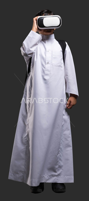 إيماءات التفاعل والاندماج، تطورأساليب التعليم باستخدام التقنيات الحديثة ومحاكاة العالم الخيالي، بورتريه لولد عربي خليجي سعودي يرتدي الثوب التقليدي ويحمل الحقيبة المدرسية يلبس نظارة الواقع الإفتراضي ثلاثية الأبعاد، صورة الجسم بالطول الكامل, خلفية سوداء