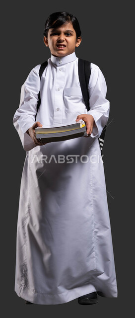 تقدم المناهج التعليمية في المملكة, إيماءات الغضب والعبوس من كثرة الواجبات المنزلية, النظر إلى الكاميرا, الشعور بالتعب خلال فترة الاختبارات والامتحانات, بورتريه مقرب لولد عربي سعودي خليجي يرتدي الثوب المدرسي ويحمل الحقيبة والكتب الدراسية الثقيلة, خلفية سوداء .