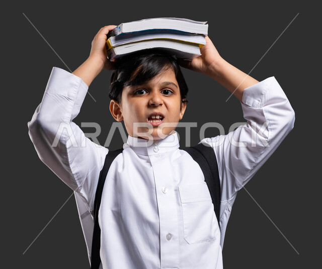 النظر إلى الكاميرا بإيماءات التعب والانزعاج من ثقل وزن المواد المدرسيه , بورتريه مقرب لولد عربي سعودي خليجي يرتدي الثوب المدرسي ويحمل الحقيبة يضع الكتب الدراسية فوق رأسه ,  بداية العام الدراسي الجديد , التعليم في المملكة العربية السعودية , خلفية سوداء .