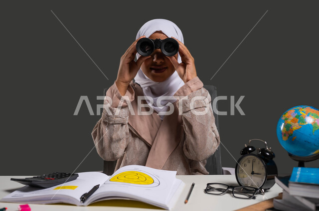 رؤية مستقبل دراسي ناجح باستخدام المنظار ثنائي العينين , بورتريه مقرب لطالبه جامعية عربية خليجية سعودية محجبة ترتدي العباءة وتجلس على كرسي مريح تبتسم وتنظر إلى الكاميرا من خلال الناظور , التخطيط المستقبلي للأهداف والطموحات ,  خلفية سوداء .