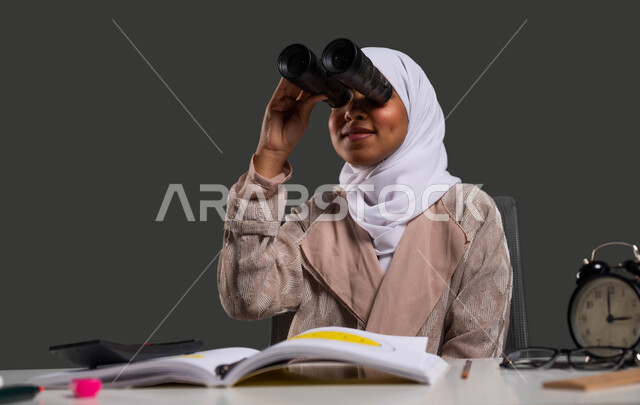 التطلع للطموحات والأهداف المستقبلية باستخدام المنظار ثنائي العينين , بورتريه مقرب لطالبة ثانوية عربية خليجية سعودية مبتسمة محجبة ترتدي العباءة وتضع منظار الرؤية , نشاطات لتحفيز العقل والفكر , إيماءات الاستمتاع والسرور ,  خلفية سوداء .