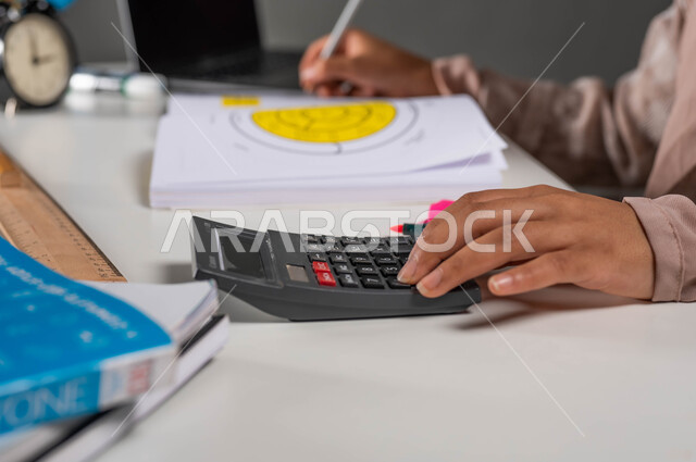 استخدام الآلة الحاسبة لإجراء العمليات الحسابية, بورتريه مقرب ليد شابه عربية خليجية سعودية تقوم بتحضير المشاريع الجامعية , أهمية التعليم في بناء مستقبل مشرق للمجتمع السعودي ,إيماءات الإجتهاد والاهتمام بالعلم والتعلم ,   خلفية سوداء .