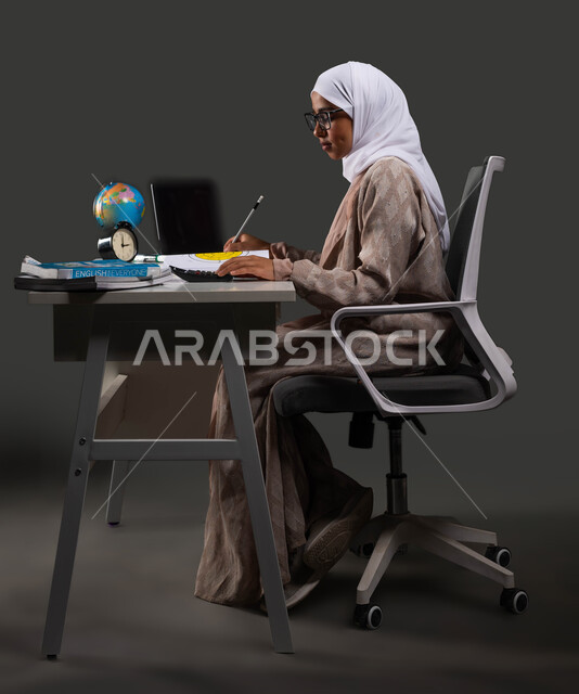 طالبة ثانوية تقوم بالتحضير للاختبارات النهائية, بورتريه من الجانب لشابه عربية خليجية سعودية محجبة ترتدي العباءة والنظارة الطبية وتجلس على كرسي مريح ,مفهوم التعليم المنزلي عبر الانترنت ,إيماءات الإجتهاد والاهتمام بالعلم والتعلم ,   خلفية سوداء .