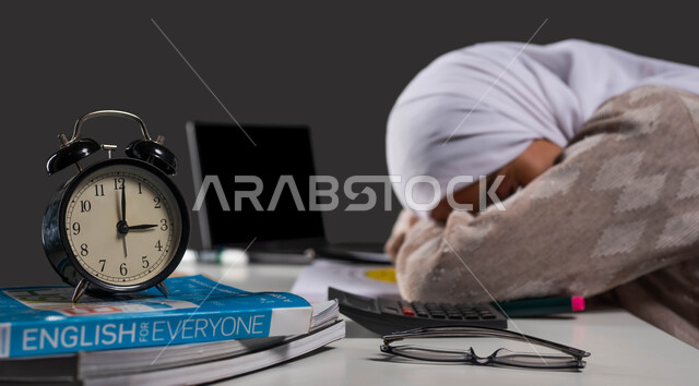انتهاء الدراسة ودخول وقت النوم اثناء مراجعة الدروس اليومية، بورتريه مقرب من الجانب لطالبة جامعية عربية سعودية خليجية ترتدي عباءة ملونة نائمة على طاولة الدراسة،أهمية الراحة اثناء المذاكره، خلفية سوداء