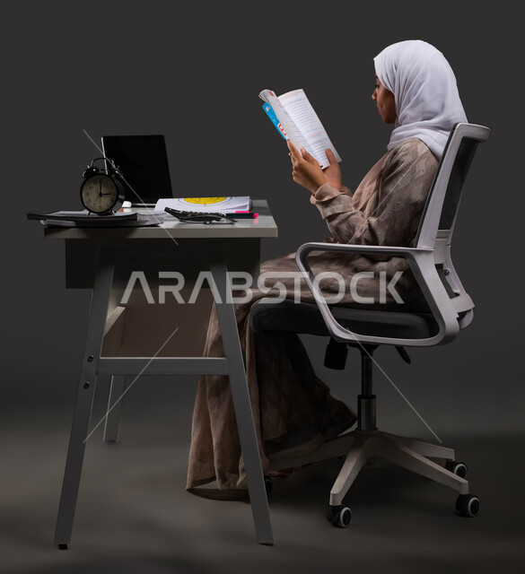 مراجعة الدروس اليومية في المنزل لتعزيز فهم المعلومات، بورتريه لطالبة ثانوي عربية سعودية خليجية ترتدي عباءة ملونة تقرأ من كتابها المدرسي، المقررات والمناهج التعليمية الحديثة والمطورة في السعودية، خلفية سوداء