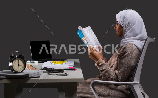 جودة ومستقبل التعليم في مدارس المملكة, مذاكرة ومتابعة المقررات المدرسية، بورتريه من الجانب لطالبة جامعية عربية سعودية خليجية ترتدي عباءة ملونة تجلس على طاولة الدراسة وتراجع دروسها اليومية، خلفية سوداء