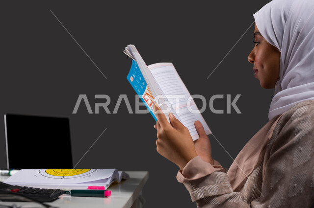 مذاكرة ومتابعة المقررات المدرسية، بورتريه مقرب من الجانب  لطالبة جامعية عربية سعودية خليجية ترتدي عباءة ملونة تجلس على طاولة الدراسة وتراجع دروسها اليومية، جودة ومستقبل التعليم في مدارس المملكة، خلفية سوداء