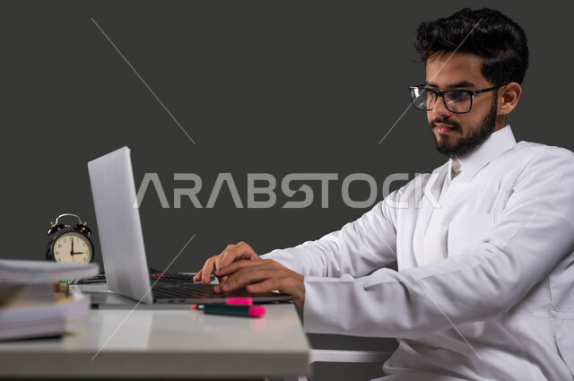 متابعة الحصص الدراسية عبر الاونلاين، بورتريه مقرب لطالب جامعي عربي خليجي سعودي يرتدي الزي التقليدي يستخدم الحاسب المحمول، دور التكنولوجيا في تغيير أساليب التعليم التقليدية، السعي للتفوق والنجاح، خلفية سوداء