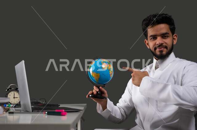 Expanding horizons and understanding cultural and natural diversity, using digital programs and applications in distance learning via laptop, interest in geographical sciences, portrait of a smiling Saudi Gulf Arab high school student wearing traditional clothing and pointing with his hand to a globe, black background.