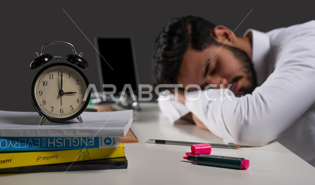 انتهاء وقت الدراسة ودخول وقت النوم, ايماءات التعب والاجهاد اثناء المذاكره، السعي للتفوق والنجاح، بورتريه مقرب لطالب جامعي عربي خليجي سعودي يرتدي الزي التقليدي نائم على طاولة الدراسة، التعليم في المملكة العربية السعودية، خلفية سوداء.