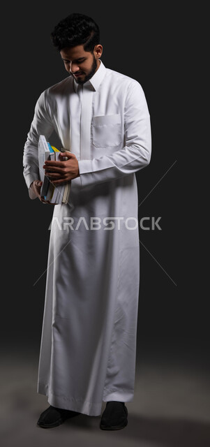 دور المدارس في تعزيز القيم والأخلاق لدى الطلاب، السعي للتفوق والنجاح، بورتريه مقرب لطالب ثانوي عربي خليجي سعودي مبتسم يرتدي الزي التقليدي وينظرالى كتبه المدرسية،التعليم في المملكة العربية السعودية، صورة الجسم بالطول الكامل، خلفية سوداء.