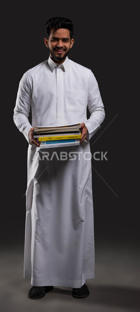 أهمية المدارس في توفير بيئة تعليمية آمنة ومحفزة للطلاب، السعي للتفوق والنجاح، بورتريه لطالب ثانوي عربي خليجي سعودي مبتسم يرتدي الزي التقليدي ويحمل كتبه المدرسية، العلم مفتاح التطور والنجاح، صورة الجسم بالطول الكامل، خلفية سوداء.