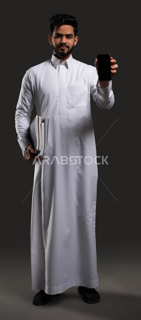 The role of technology in improving student performance, the importance of education in developing Saudi society, portrait of a Saudi Gulf Arab high school student wearing traditional clothing and holding a mobile phone in his hand with a blank black screen, full-length body photo, black background.