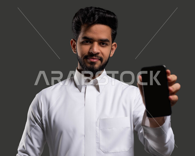 دور التكنولوجيا في تحسين تجربة التعلم، أهمية التعليم في تطوير المجتمع السعودي، بورتريه مقرب لطالب جامعي عربي خليجي سعودي يرتدي الزي التقليدي ويمسك بيده الهاتف المحمول بشاشة سوداء فارغة، خلفية سوداء.