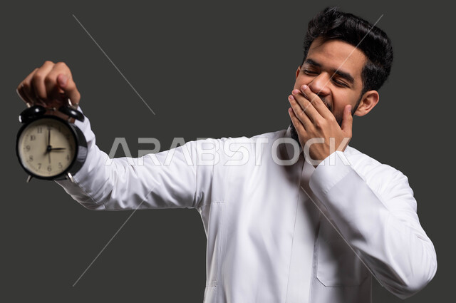 The difficulty of waking up early for school, developing curricula and educational materials in Saudi Arabia, the importance of education in developing Saudi society, a close-up portrait of a Saudi Gulf Arab high school student wearing traditional clothing and holding an alarm clock in his hand, black background.