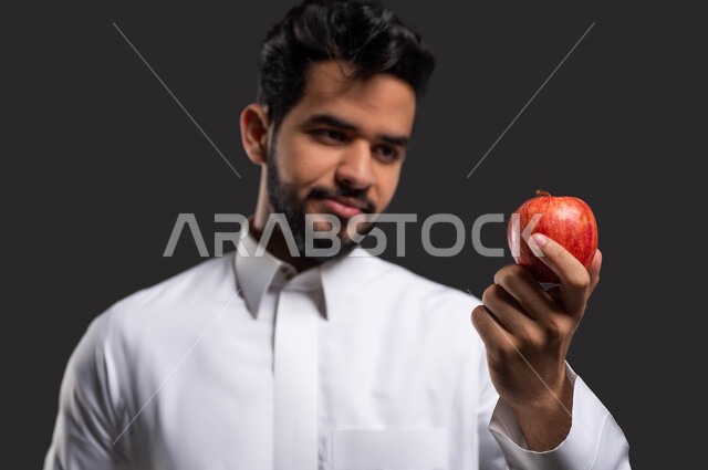 التشجيع على تناول الطعام الصحي ، تطوير المناهج والمواد التعليمية في السعوديه،أهمية التعليم في تطوير المجتمع السعودي، بورتريه مقرب لطالب ثانوي عربي خليجي سعودي يرتدي الزي التقليدي ويمسك بيده حبة تفاح، ايماءات الفرح بدخول وقت الفسحة، خلفية سوداء.