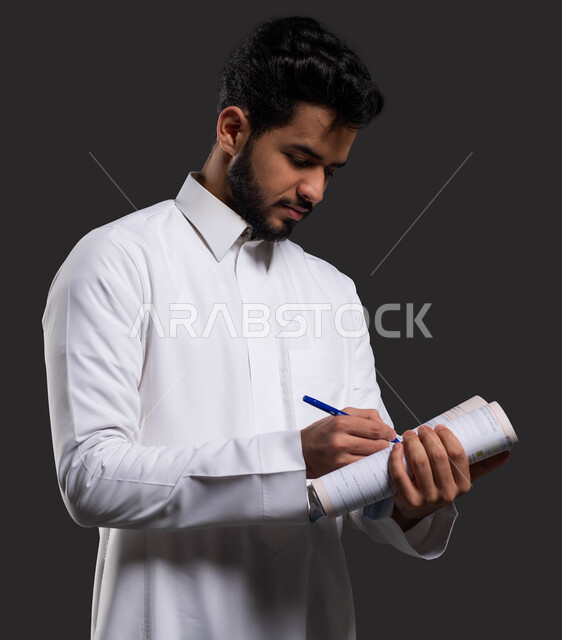 ايماءات الاندماج والتركيز في الكتابة، تطوير المناهج والمواد التعليمية في السعوديه،أهمية التعليم في تطوير المجتمع السعودي، بورتريه مقرب لطالب جامعي عربي خليجي سعودي يرتدي الزي التقليدي ويكتب على الدفتر، خلفية سوداء.