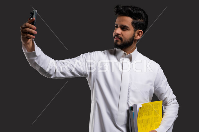 التعلم التفاعلي عبر الانترنت، بورتريه مقرب لطالب جامعي خليجي سعودي يرتدي الزي التقليدي ويأخذ صورة سيلفي بالهاتف، استخدام البرامج والتطبيقات الرقمية في التعلم، العودة الى المدارس، خلفية سوداء