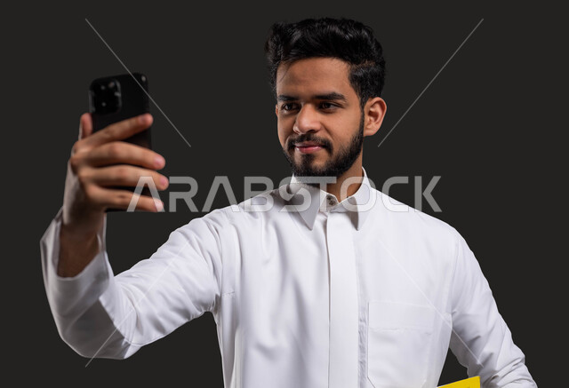 The role of technology in changing traditional education methods, a close-up portrait of a Saudi Gulf high school student wearing traditional clothing and taking a selfie with the phone, using digital programs and applications in learning, black background