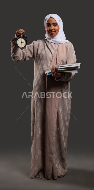 استغلال الوقت في الدراسة والمذاكرة، النظر إلى الكاميرا بإيماءات وجه تدل على السرور، أهمية التعليم في المجتمع السعودي, بورتريه لطالبة جامعية عربية خليجية سعودية محجبة ترتدي العباءة وتحمل بيدها ساعة منبه, صورة الجسم بالكامل، خلفية سوداء.