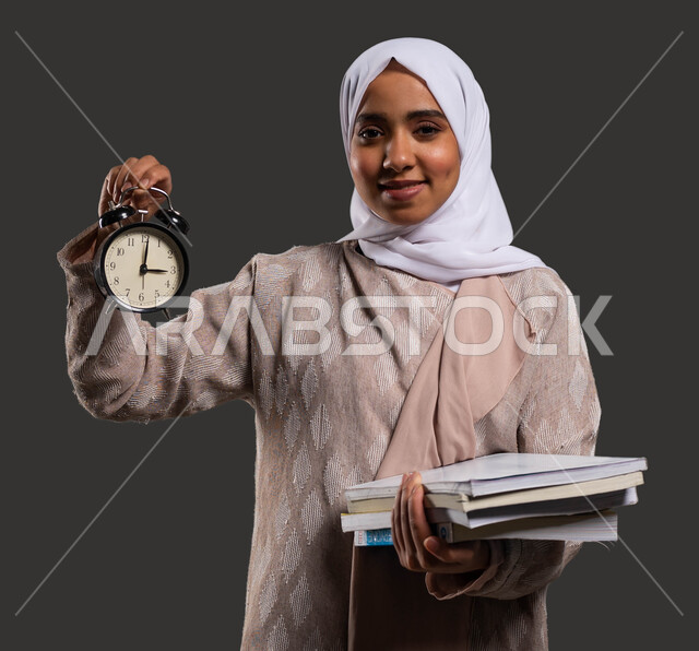 النظر إلى الكاميرا بإيماءات وجه تدل على السرور، استغلال الوقت في الدراسة والمذاكرة، أهمية التعليم في المجتمع السعودي, بورتريه مقرب لطالبة جامعية عربية خليجية سعودية محجبة ترتدي العباءة وتحمل بيدها ساعة منبه, خلفية سوداء.