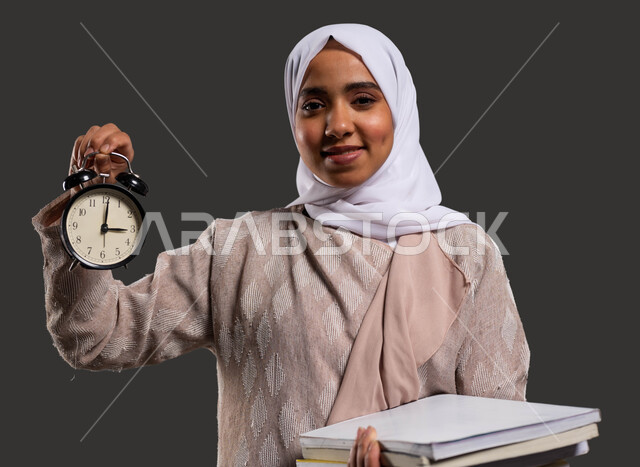 استغلال الوقت في الدراسة والمذاكرة، النظر إلى الكاميرا بإيماءات وجه تدل على السرور، أهمية التعليم في المجتمع السعودي, بورتريه مقرب لطالبة جامعية عربية خليجية سعودية محجبة ترتدي العباءة وتحمل بيدها ساعة منبه, خلفية سوداء.