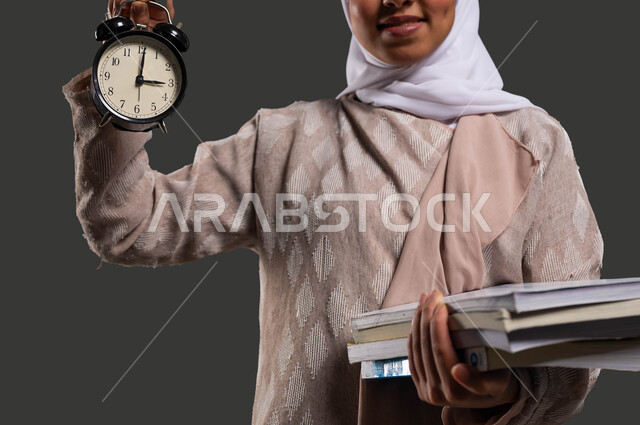 النظر إلى الكاميرا بإيماءات وجه تدل على السرور، استغلال الوقت في الدراسة والمذاكرة، أهمية التعليم في المجتمع السعودي, بورتريه مقرب لطالبة جامعية عربية خليجية سعودية محجبة ترتدي العباءة وتحمل بيدها ساعة منبه, خلفية سوداء.