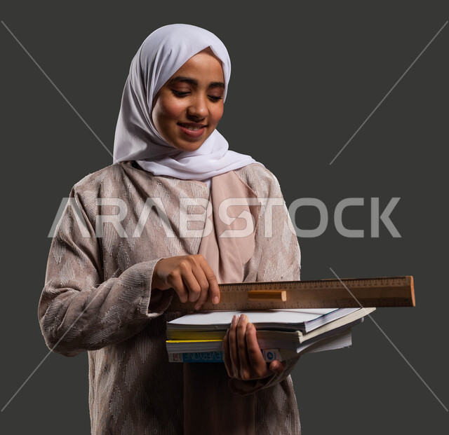 قياس المسافات ورسم الخطوط المستقيمة بالمسطرة الخشبية، دور المدارس في تنمية القيم والأخلاق الإنسانية لدى الطلاب، بورتريه مقرب لطالبة ثانوي سعودية ترتدي عباءة ملونة وتحمل بيدها الكتب المدرسية، أهمية التعليم في بناء مستقبل مشرق للمجتمع السعودي، خلفية سوداء.