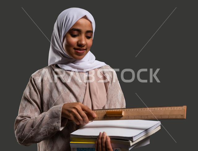 قياس المسافات ورسم الخطوط المستقيمة بالمسطرة الخشبية، دور المدارس في تنمية القيم والأخلاق الإنسانية لدى الطلاب، بورتريه مقرب لطالبة ثانوي سعودية ترتدي عباءة ملونة وتحمل بيدها الكتب المدرسية، أهمية التعليم في بناء مستقبل مشرق للمجتمع السعودي، خلفية سوداء.