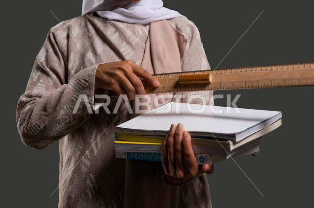 قياس المسافات ورسم الخطوط المستقيمة بالمسطرة الخشبية، دور المدارس في تنمية القيم والأخلاق الإنسانية لدى الطلاب، بورتريه مقرب لطالبة ثانوي سعودية ترتدي عباءة ملونة وتحمل بيدها الكتب المدرسية، أهمية التعليم في بناء مستقبل مشرق للمجتمع السعودي، خلفية سوداء.