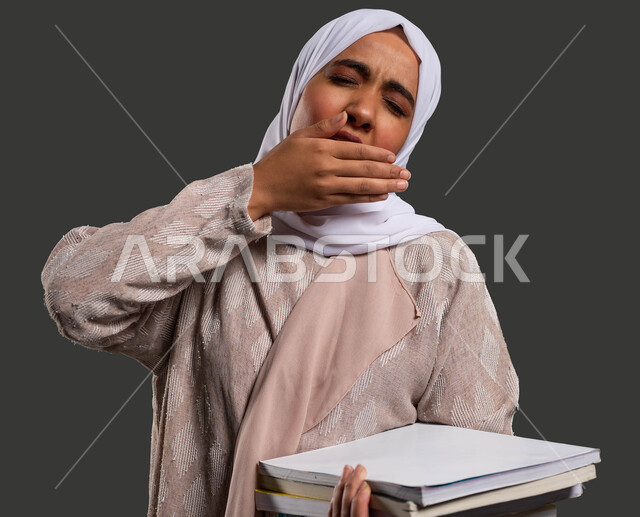 Feeling tired, lethargic, and exhausted while studying, difficulty waking up early, the importance of education in Saudi society, close-up portrait of a veiled Saudi Gulf Arab university student wearing an abaya and placing her hand over her mouth with sleepy gestures, black background.