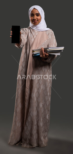 The role of technology in improving student performance, the importance of education in developing Saudi society, a portrait of a Saudi Gulf Arab high school student wearing an abaya and holding a mobile phone in her hand with a blank black screen, full-length body photo, black background.
