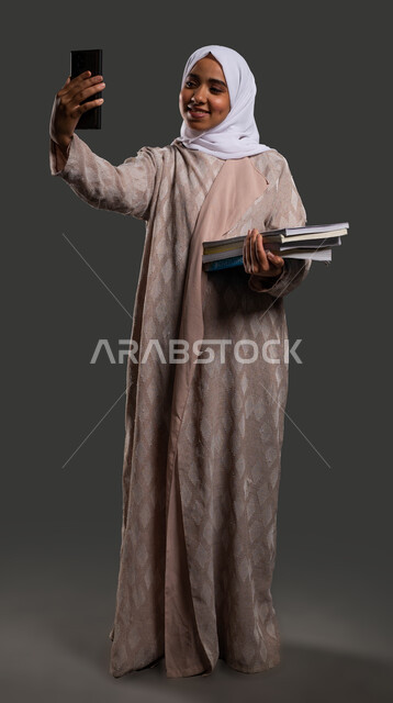 استخدام البرامج والتطبيقات الرقمية في التعلم، مفهوم التعلم التفاعلي عبر الانترنت، بورتريه لطالبة جامعية عربية خليجية سعودية محجبة مبتسمة ترتدي العباءة وتأخذ صورة سيلفي بالهاتف، العودة إلى المدارس في السعودية، صورة الجسم بالكامل، خلفية سوداء.