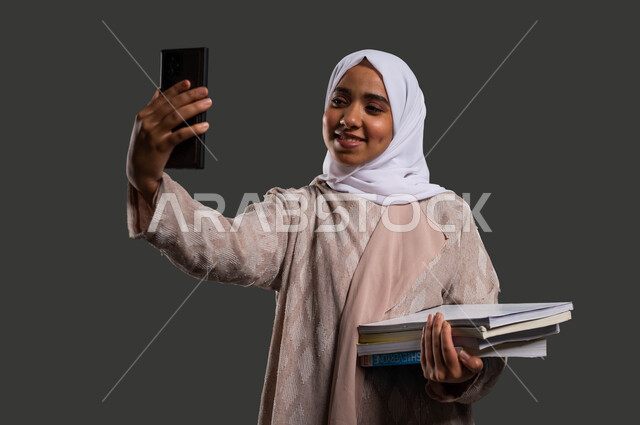 The concept of interactive online learning, a close-up portrait of a veiled Saudi Arabian Gulf university student, smiling, wearing an abaya and taking a selfie with the phone, using digital programs and applications in learning, returning to schools in Saudi Arabia, black background.