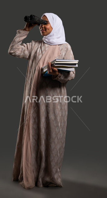 رؤية مستقبليه لتحقيق النجاح والتفوق الدراسي, بورتريه لطالبة جامعية عربية خليجية سعودية محجبة ترتدي العباءة وتنظر من خلال المنظار, استخدام الناظور ثنائي العينين لاكتشاف الأشياء البعيدة, صورة الجسم بالكامل، خلفية سوداء .