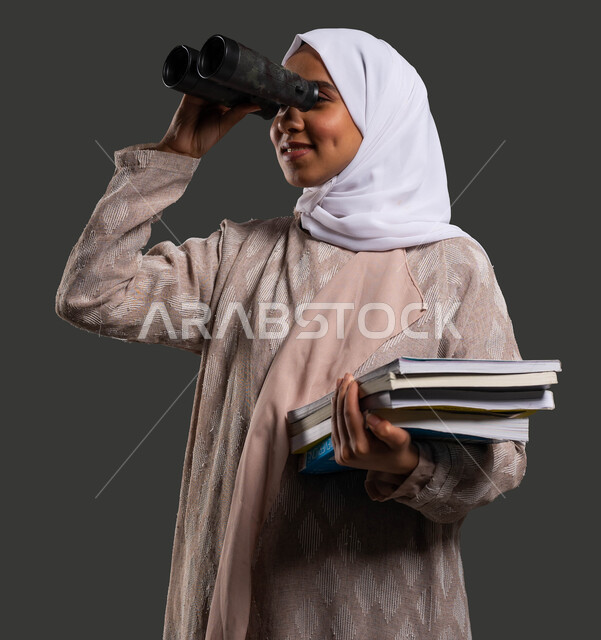 Looking forward to future ambitions and goals using binoculars, activities to stimulate the mind and thought, a close-up portrait of a smiling, veiled Saudi Arabian Gulf high school student wearing an abaya and wearing binoculars, gestures of enjoyment and pleasure, a black background.