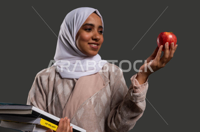 دور المدارس في رفع مستوى الوعي لدى الطلاب، تحسين مستوى الذكاء بتناول الطعام الصحي، أهمية التعليم في تطوير المجتمع السعودي، بورتريه مقرب لطالبة ثانوية عربية خليجية سعودية محجبة مبتسمة ترتدي العباءة وتمسك بيديها حبة تفاح وكتبها المدرسية، خلفية سوداء.