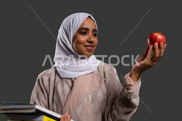 Improving the level of intelligence by eating healthy food, developing curricula and educational materials in Saudi Arabia, the importance of education in developing Saudi society, a close-up portrait of a Saudi Gulf Arab high school student wearing an abaya, holding an apple and her school books in her hands, black background.