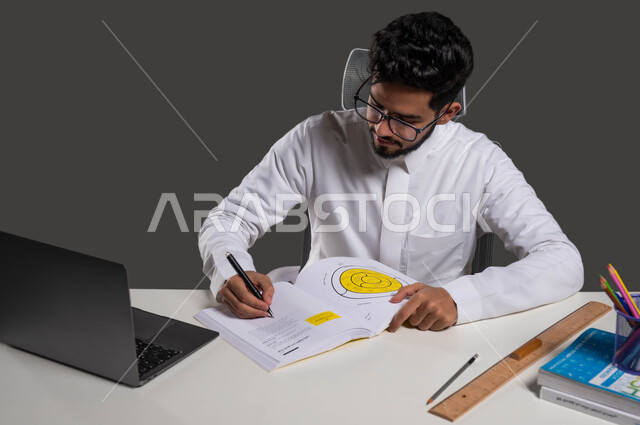 تطوير المناهج التعليمية في السعوديه، متابعة الدروس والواجبات عبر الانترنت، أهمية التعليم في تطوير المجتمع السعودي، بورتريه لطالب جامعي عربي سعودي يرتدي الزي التقليدي والنظارة الطبية يجلس أمام الطاولة الدراسية وجهاز الحاسوب ويقوم بكتابة واجباته، خلفية سوداء.